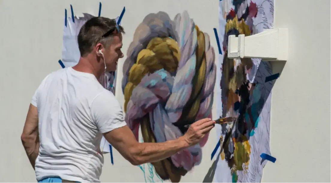 Man wearing a white t-shirt painting a colorful abstract design on a white wall outdoors.