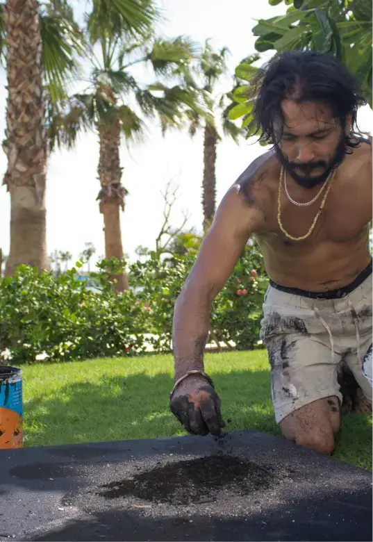 Shirtless man with dirty hands kneeling on grass, working with black paint or ink on a flat surface outdoors near palm trees.