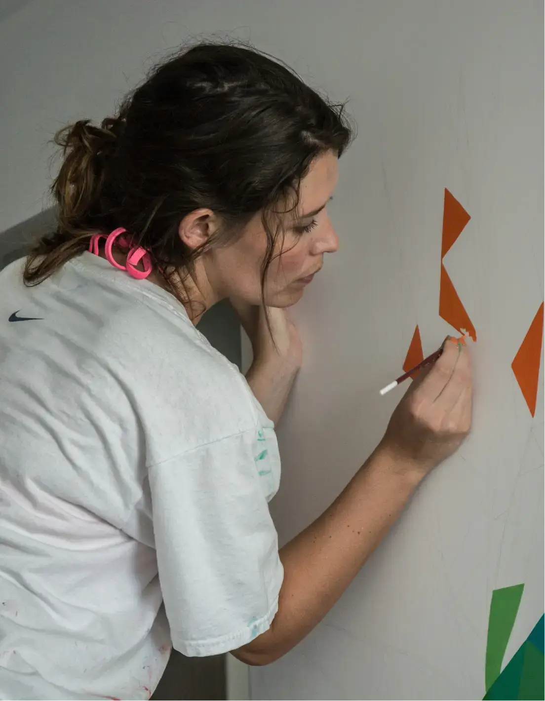 Woman with dark hair tied back, painting orange geometric shapes on a white wall.