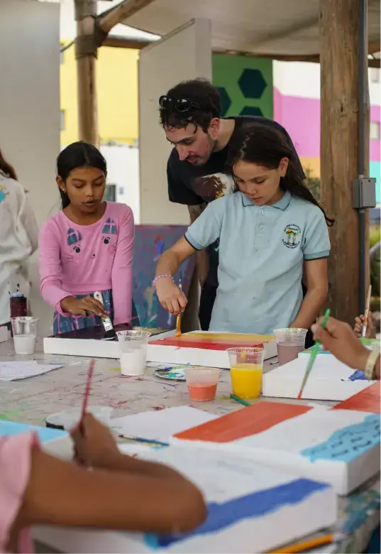 Two young girls painting canvases at a table with a man supervising closely.