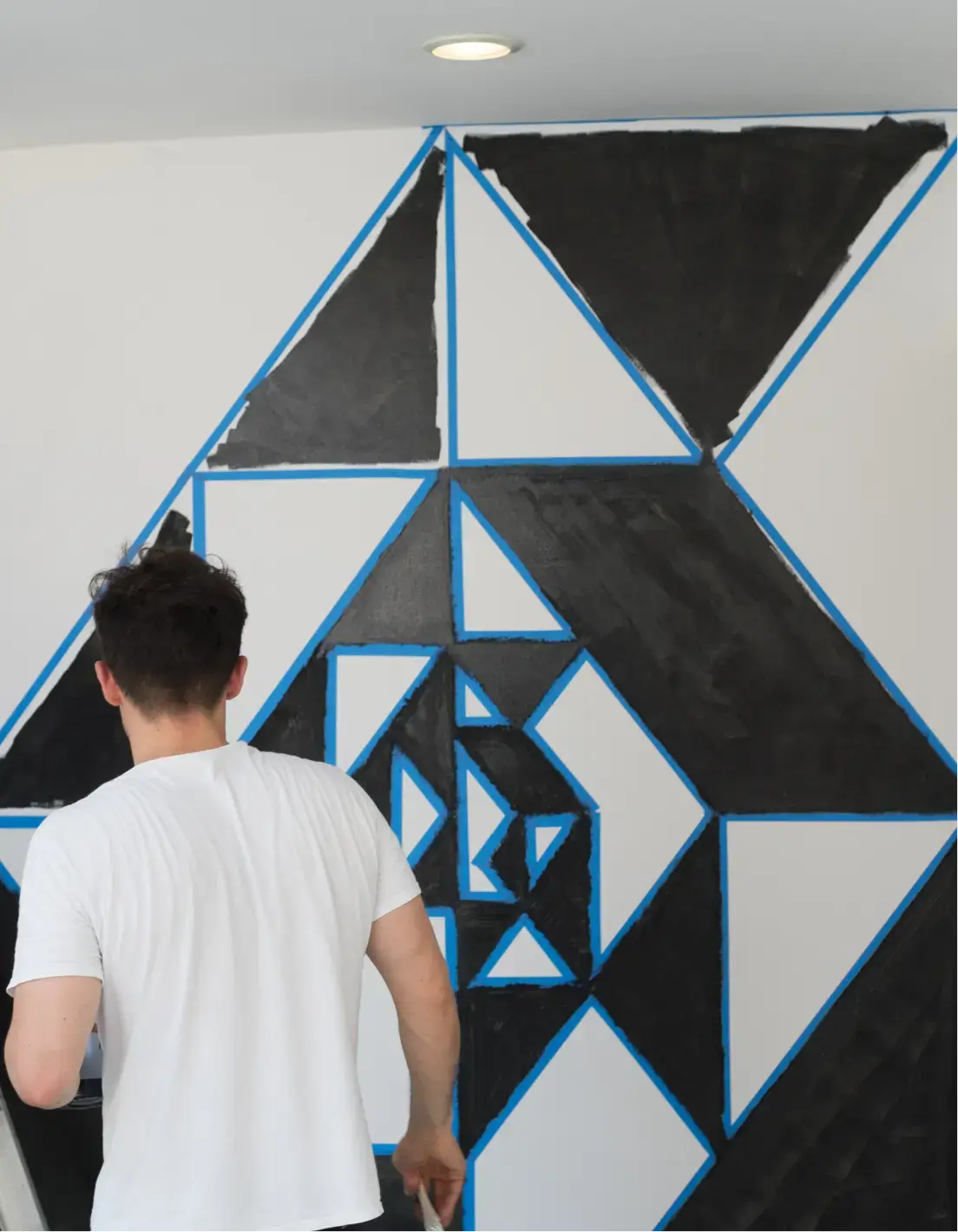 Man in a white t-shirt painting a geometric black and white triangle pattern on a wall, outlined with blue tape.