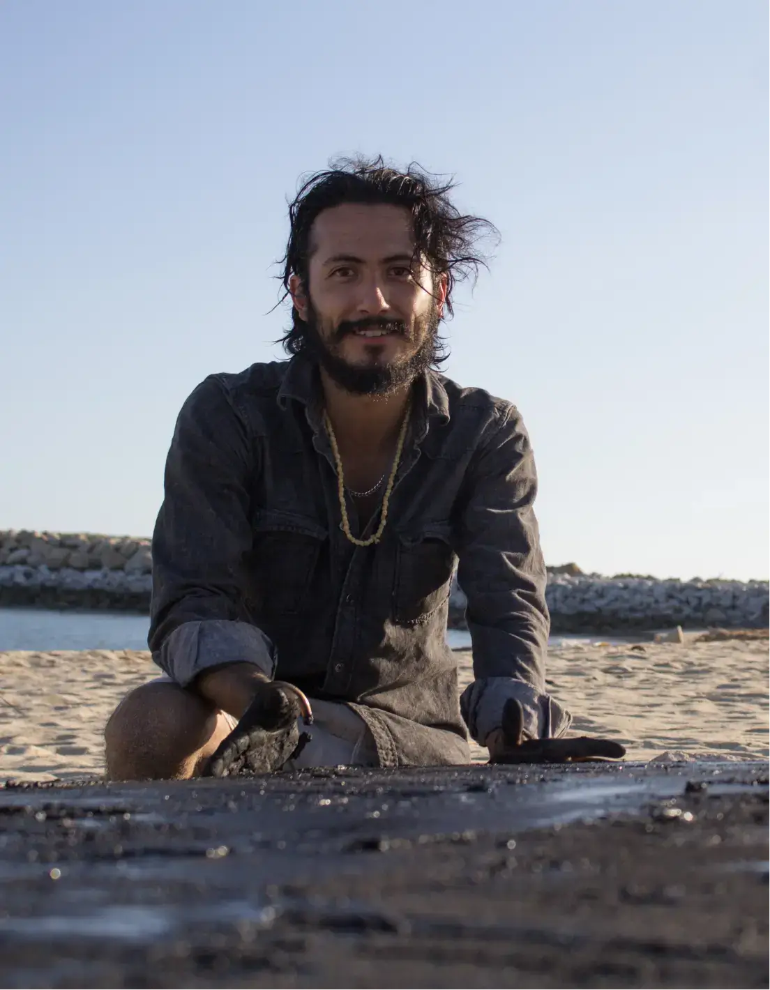 Man with dark hair and beard kneeling on a sandy beach with a stone barrier and calm water in the background.