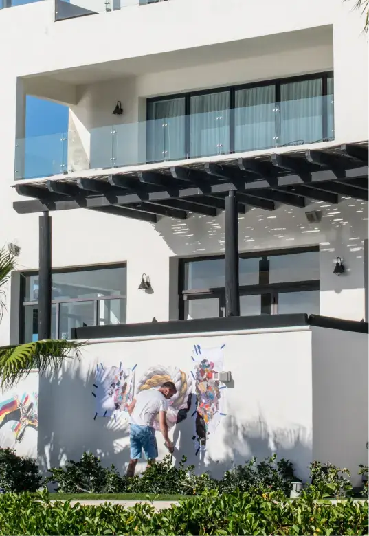 Man in white shirt and blue shorts working on a colorful mural on a white wall outside a modern white building with glass balcony and black pergola.