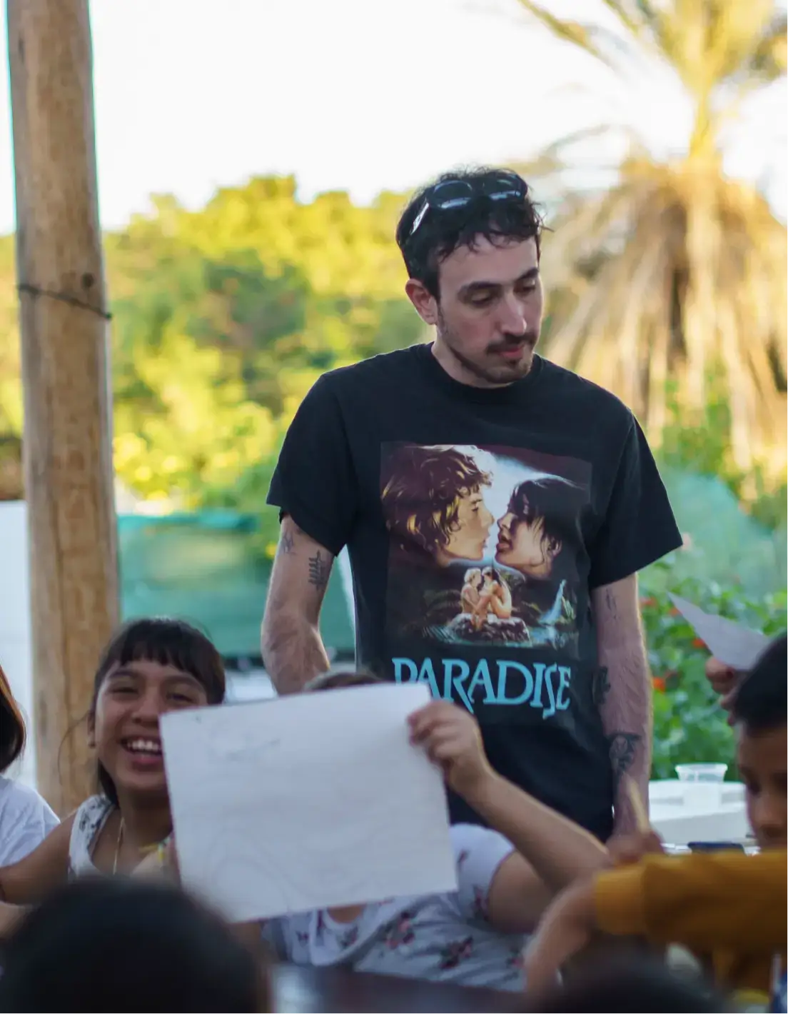 Man wearing a black 'Paradise' t-shirt looks down while children around him smile and hold up drawings outdoors.