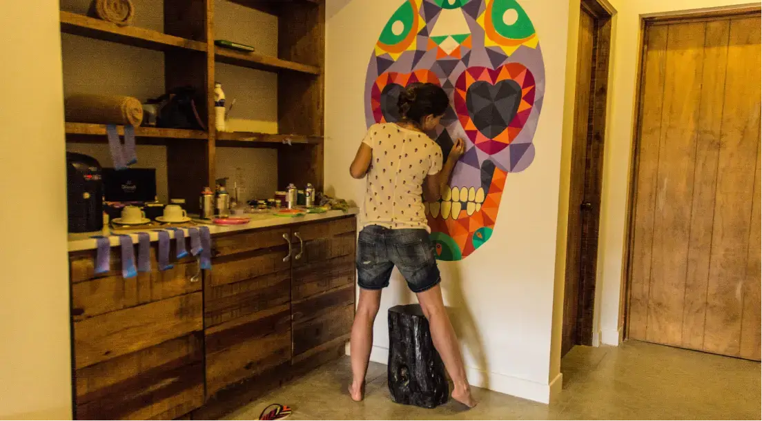 Person painting a colorful geometric skull mural on a white wall in a room with wooden cabinets.