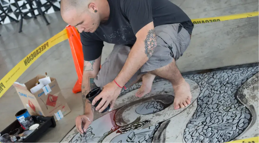 Tattooed man in a black shirt and gray shorts barefoot painting detailed black and white artwork on the floor inside a cordoned-off area.