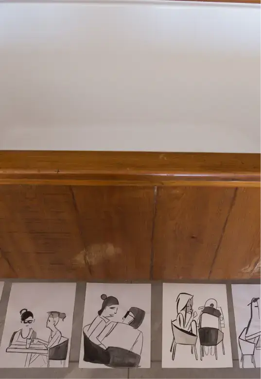 Four black-and-white minimalist drawings on the floor depict people sitting and interacting in various poses beneath a wooden piece of furniture.