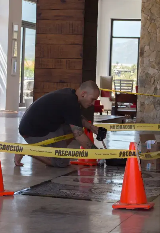 Man crouching barefoot inside a room, working near a rug surrounded by orange cones and yellow caution tape that reads 'PRECAUCIÓN.'