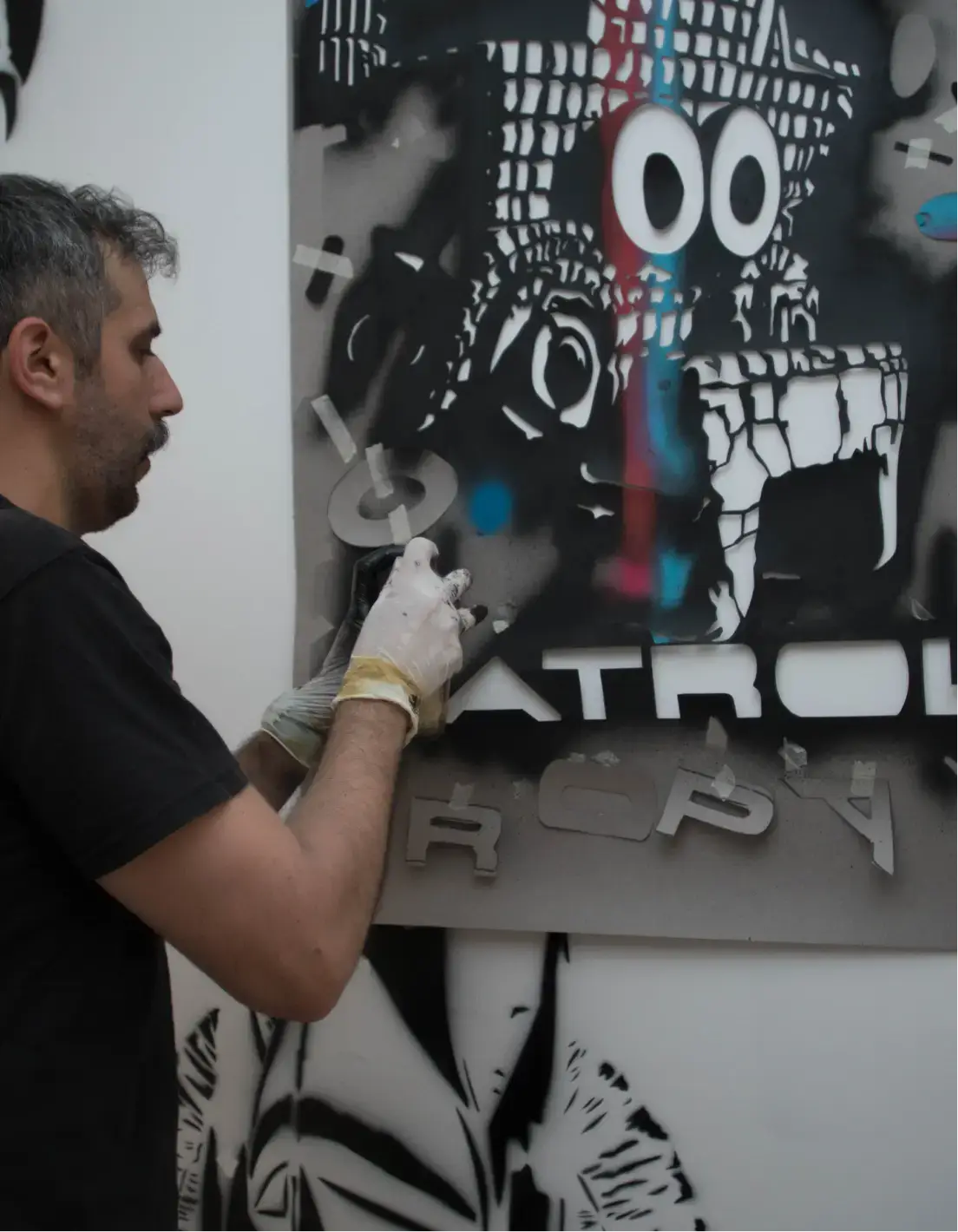 Man wearing gloves spray painting a stencil artwork with abstract face and text on a wall.