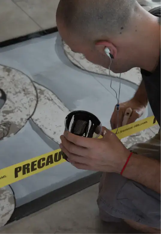 Person kneeling and painting a section of artwork while holding a paintbrush and a small container, next to yellow caution tape reading 'PRECAUTION'.