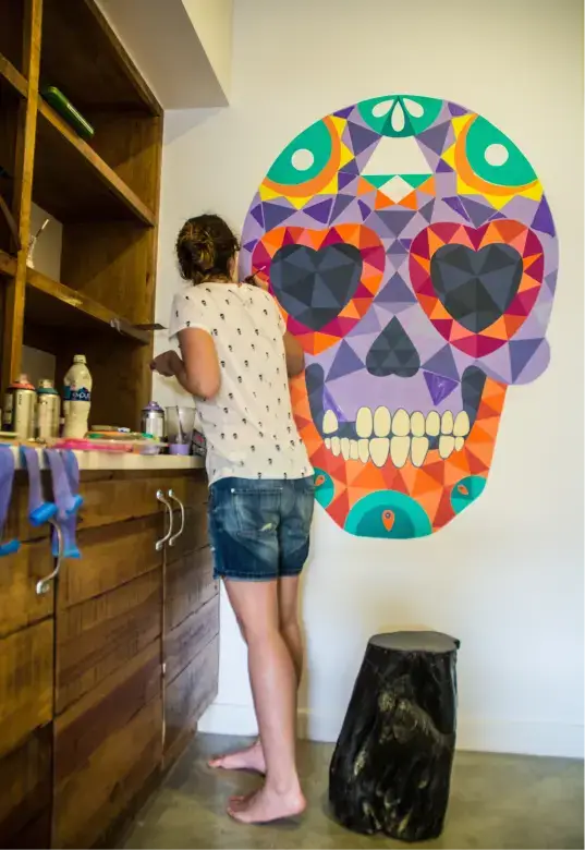 Person in a white shirt and denim shorts painting or working near a counter with a colorful geometric skull mural on the wall.