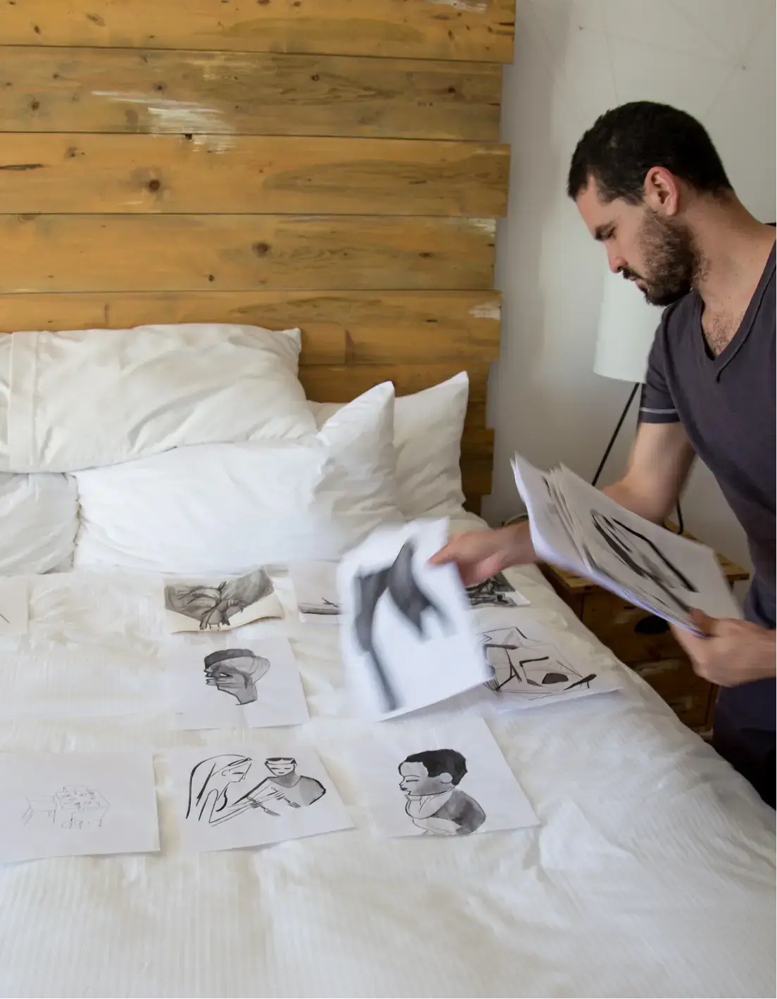 Man arranging black and white sketches on a bed with white bedding and wooden headboard.