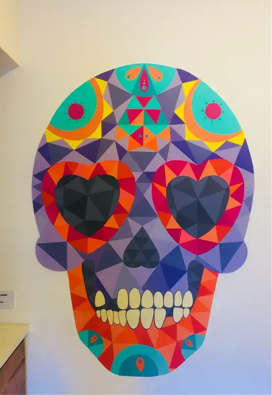 Colorful geometric skull mural with heart-shaped eyes in shades of purple, orange, red, and turquoise on a white wall.