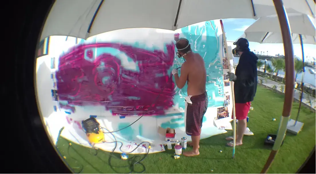 Two men creating colorful graffiti art on a large white board outdoors under umbrellas.