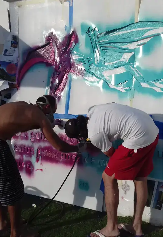 Two people wearing masks spray painting colorful dragon designs and text on a white wall outdoors.