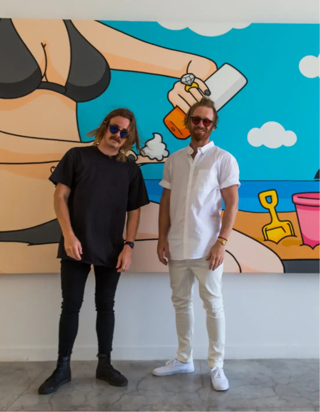 Two men wearing sunglasses standing in front of a colorful mural featuring a beach scene with a woman in a black bikini, a hand holding a cigarette, clouds, sand, and beach toys.