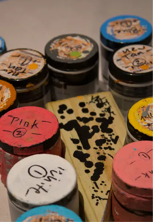 Close-up of various paint jars with handwritten labels and a wooden paint stirring paddle with black paint marks.