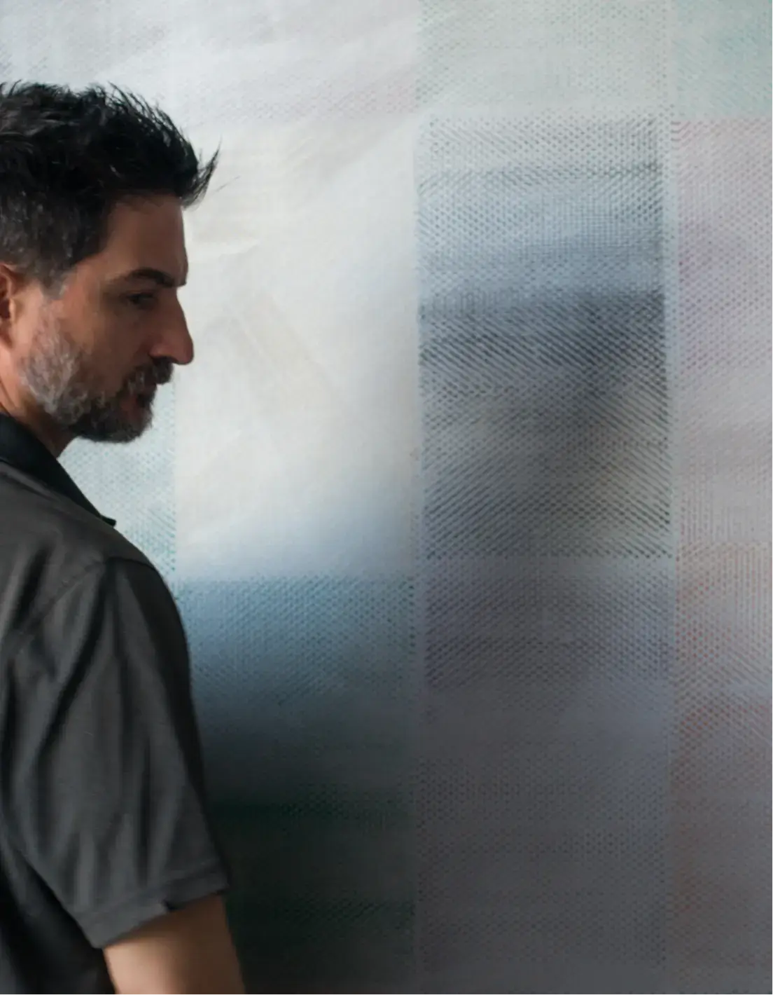 Man with dark hair and beard looking at a textured, abstract artwork on a wall.