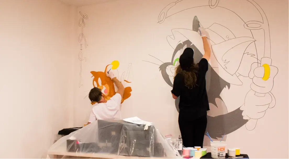 Two artists painting a wall mural featuring Tom and Jerry cartoon characters.