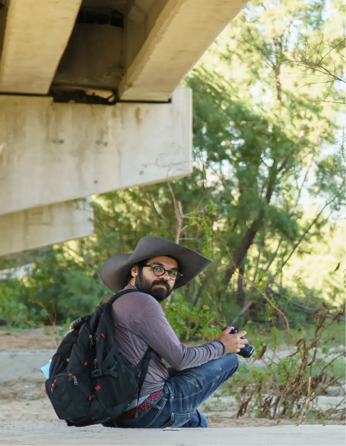 Man wearing a wide-brimmed hat and glasses sitting outdoors under a concrete structure, holding a camera and carrying a black backpack.