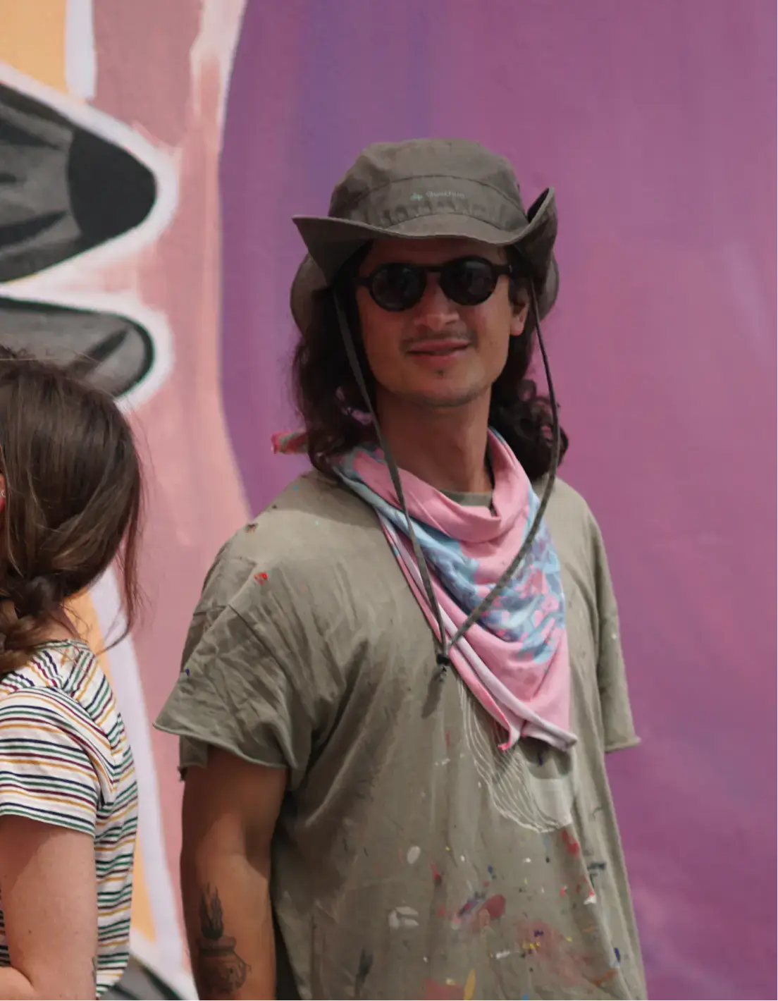 Young man with sunglasses, wide-brimmed hat, and paint-splattered shirt standing against a purple and orange mural background.