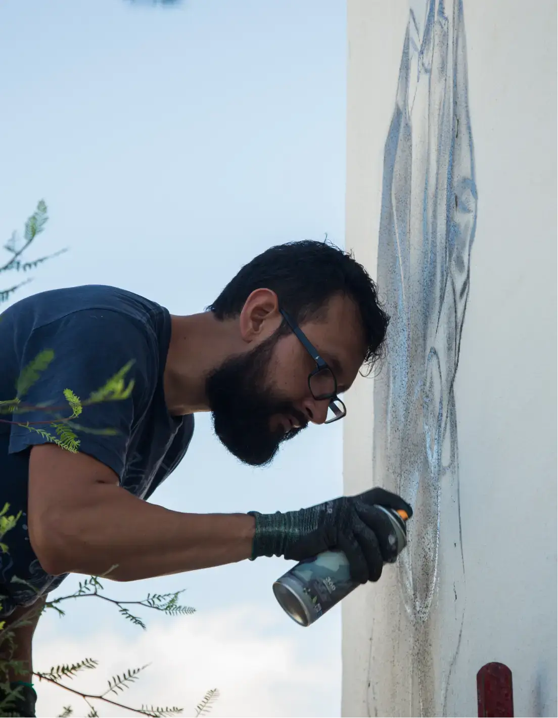 Man with glasses and a beard spray painting a mural on a white wall outdoors.