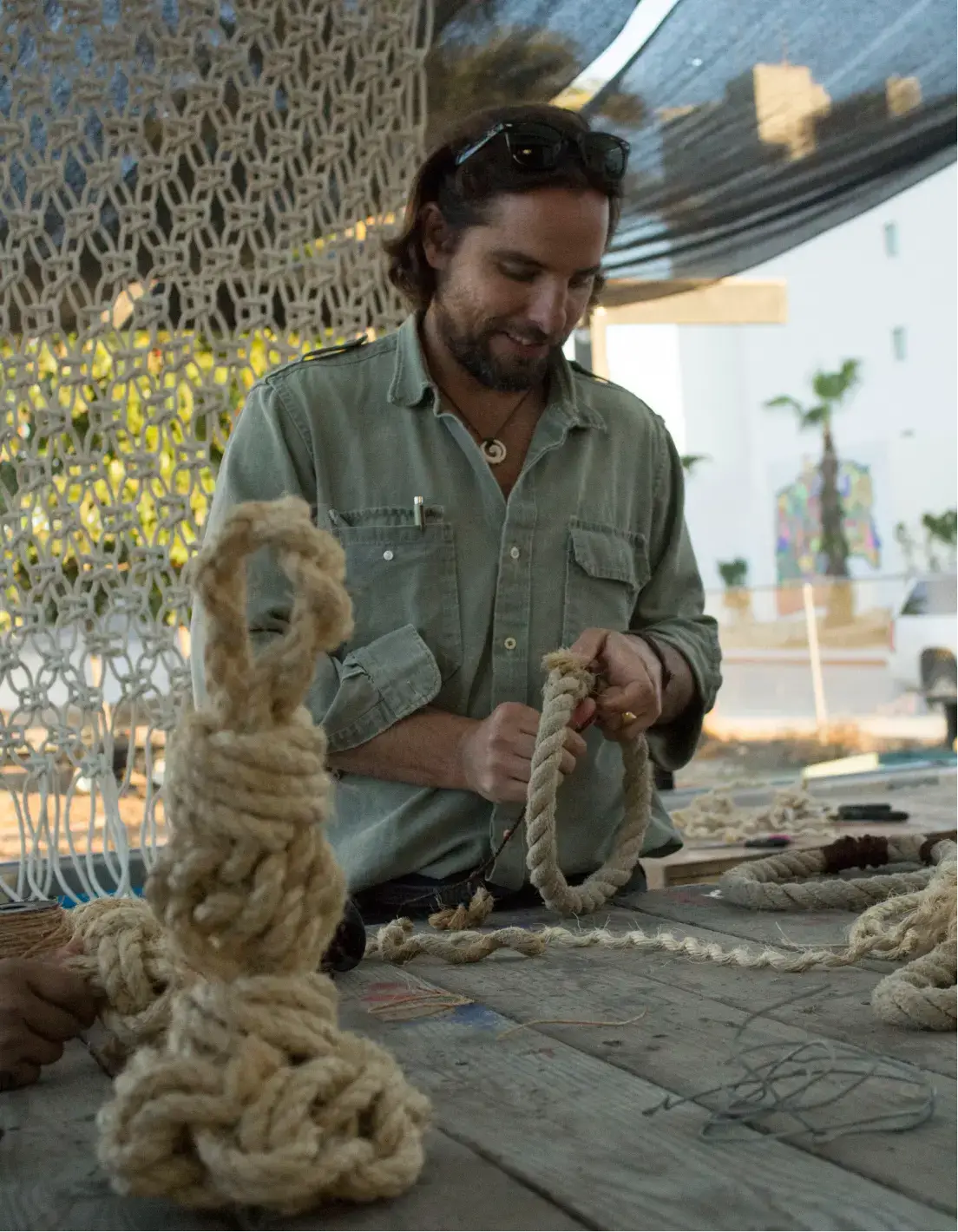 Man with beard working on crafting with thick rope at a wooden table under a shaded area.