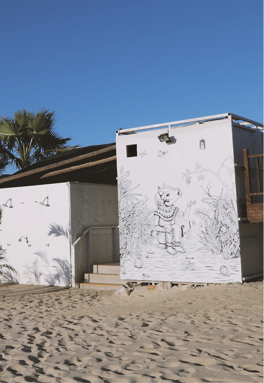 White beach building with black line drawing of an anthropomorphic animal sitting among plants and pumpkins under a clear blue sky.