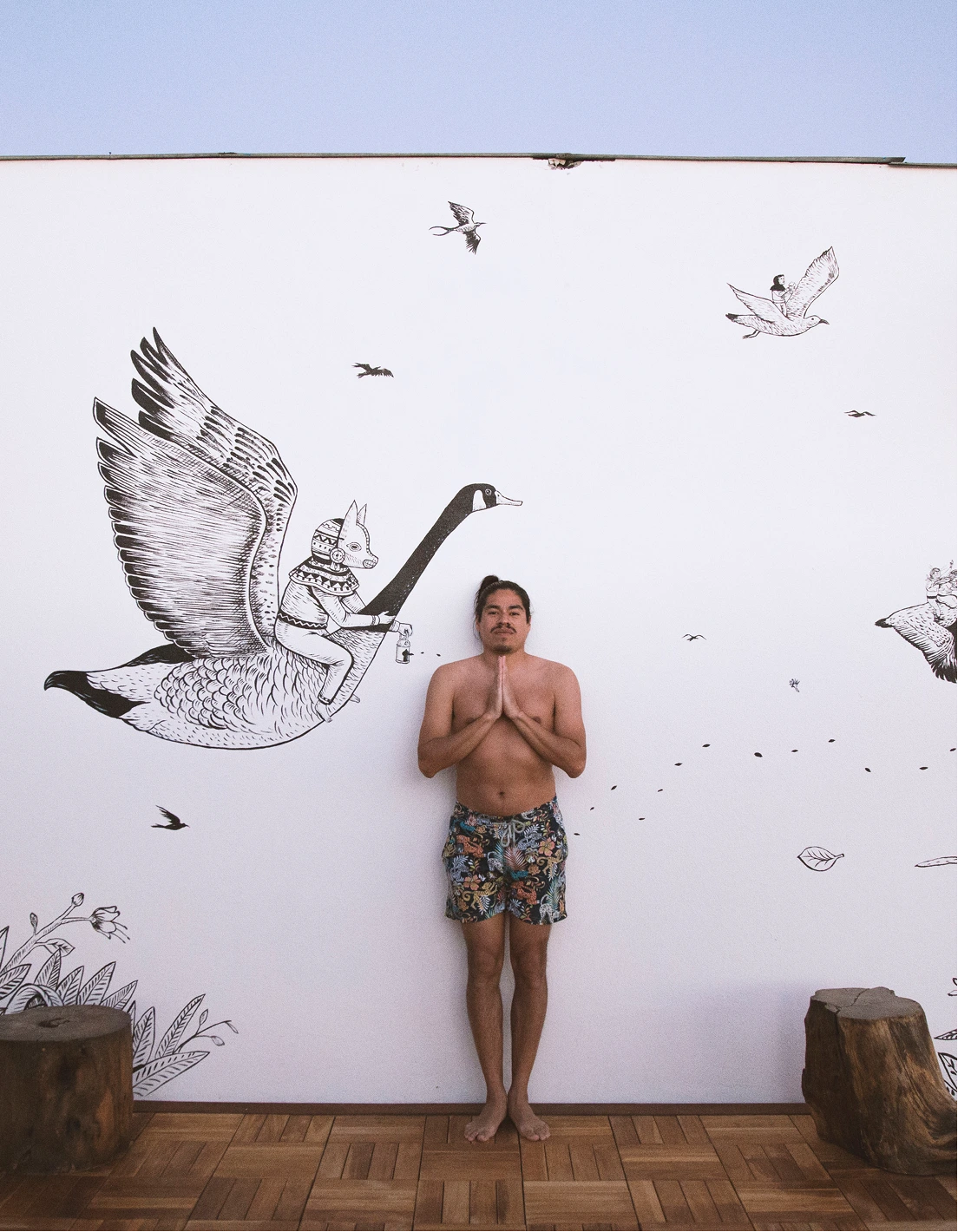 Shirtless man wearing colorful shorts stands barefoot with hands in prayer pose against a white wall mural of a mythical character riding a large flying goose.