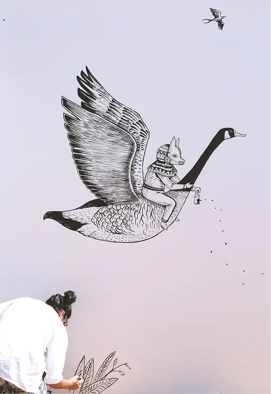 Artist painting a wall mural of a person with an animal mask riding a large flying goose holding a lantern.