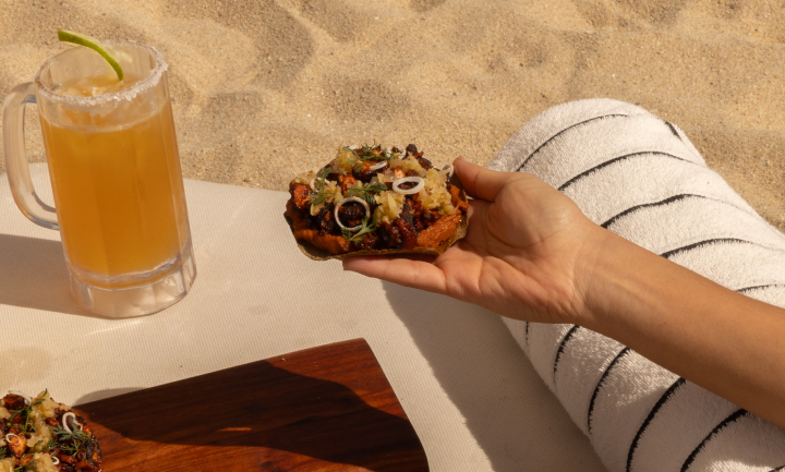 Mano sosteniendo un taco con ingredientes variados junto a una toalla a rayas sobre la arena y un vaso de cerveza con una rodaja de lima.