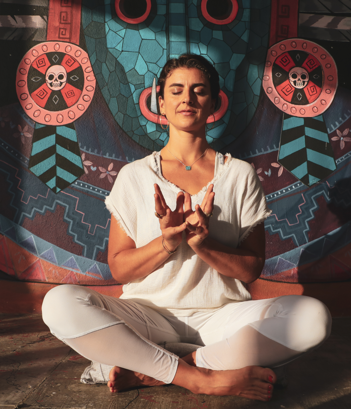 Woman in white clothes meditating cross-legged with eyes closed in front of colorful mural with skull designs.