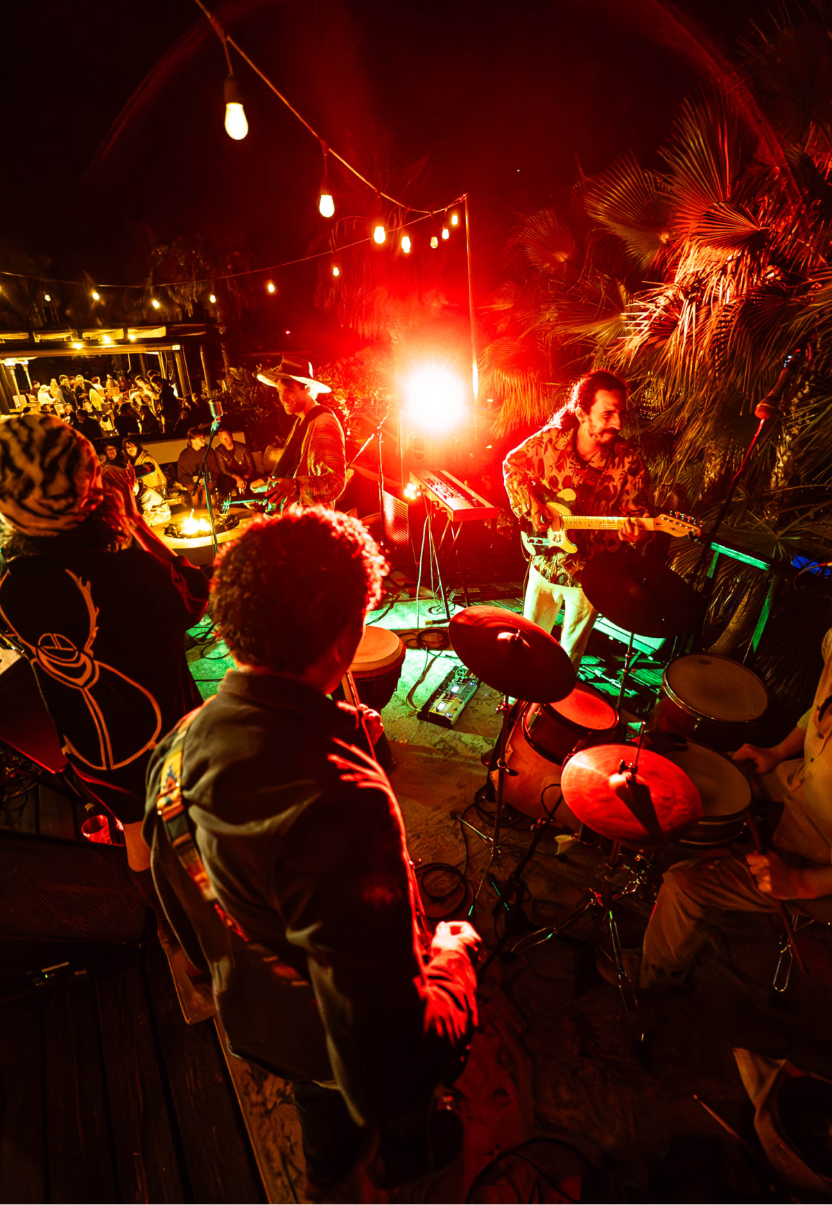 Outdoor night scene of a band playing live music with string lights overhead and an audience in the background.