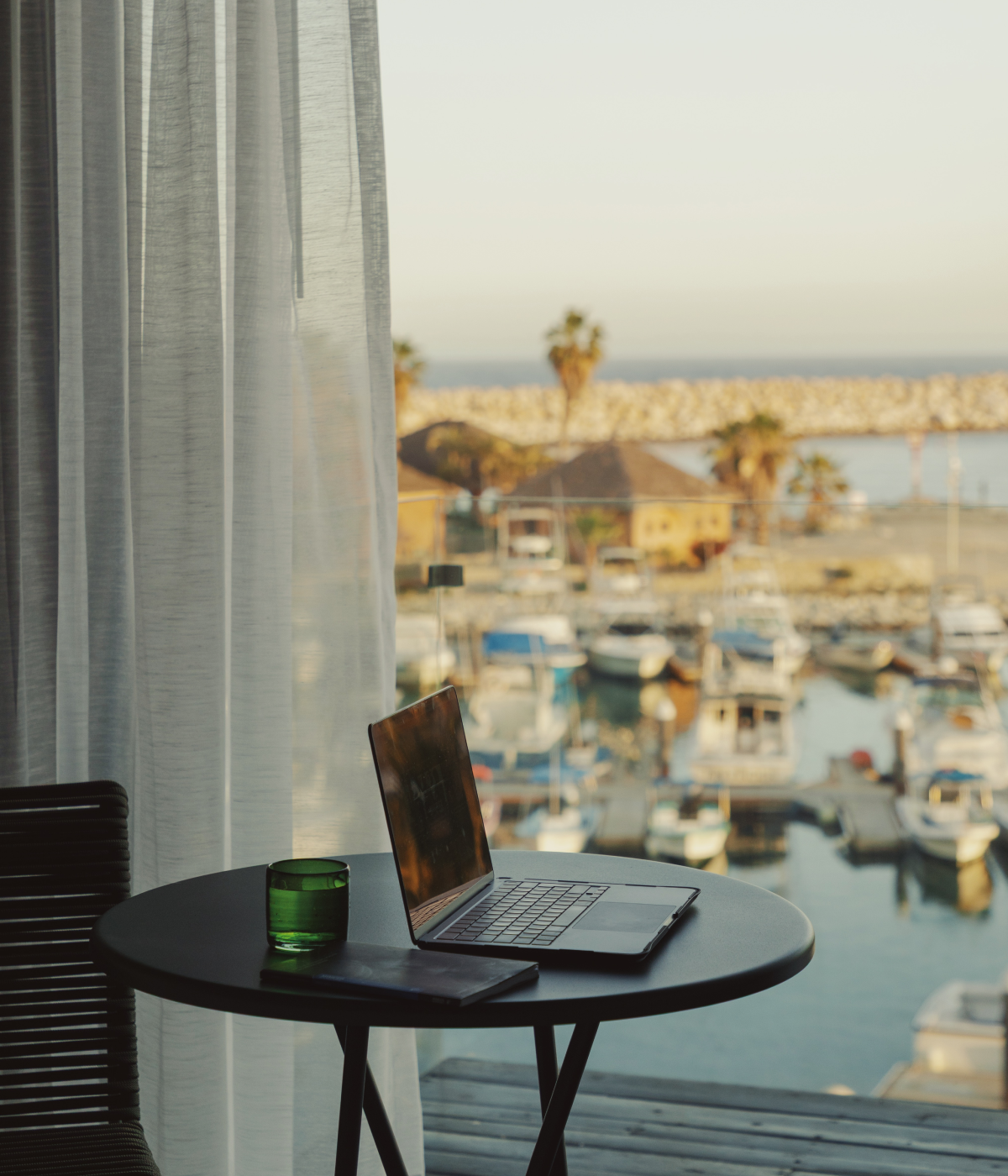 A laptop, green glass, and notebook on a round table by a window overlooking a marina with boats and palm trees.