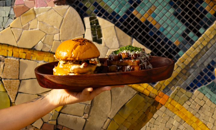 Mano sosteniendo una bandeja de madera con una hamburguesa con queso y una guarnición de patatas fritas sazonadas con hierbas y queso.