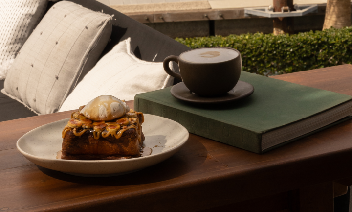 Tostada francesa cubierta con sirope y una cucharada de nata en un plato junto a una taza de capuchino sobre un libro verde, colocada sobre una mesa de madera con cojines de sofá al fondo.