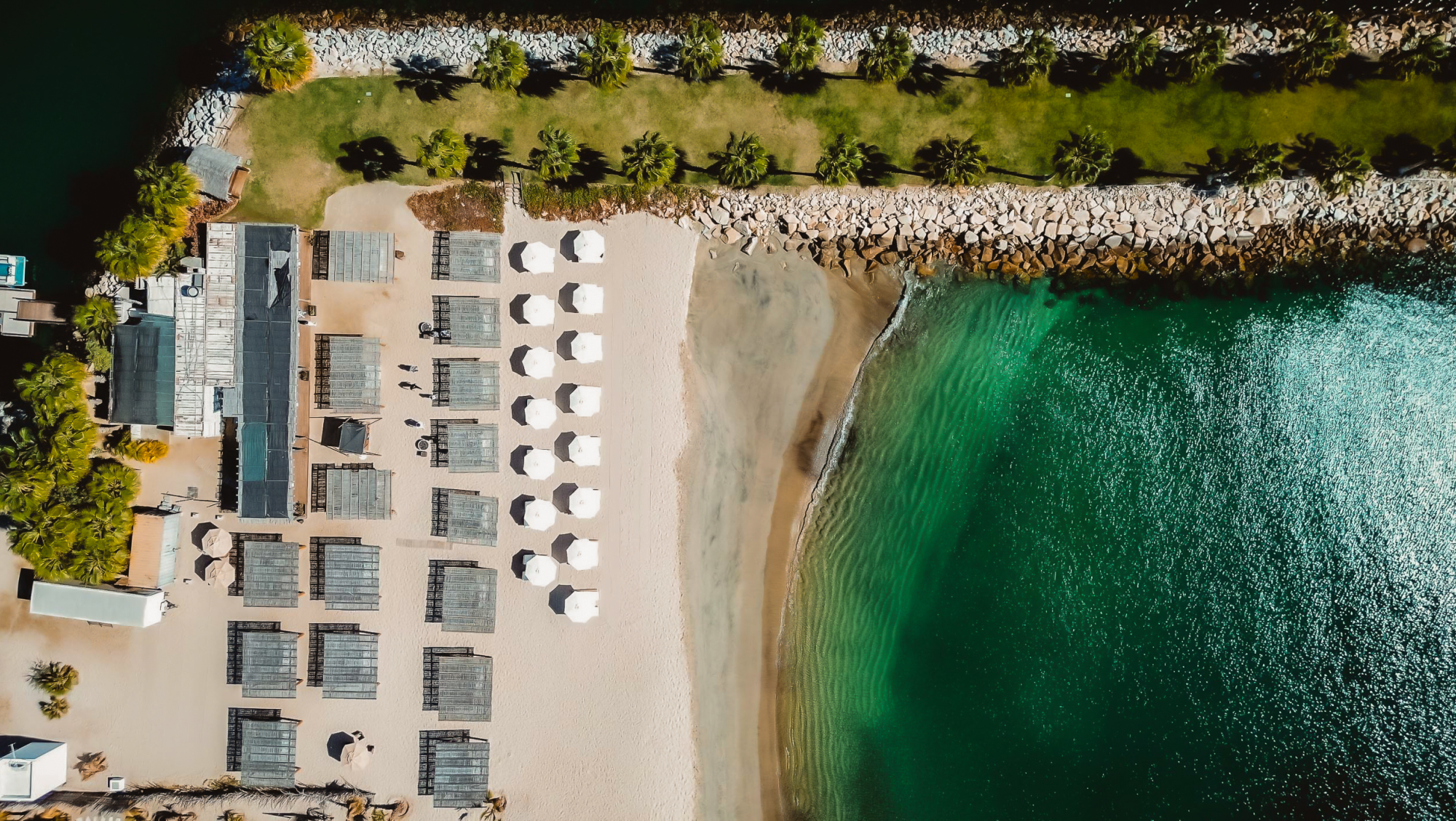 Aerial view of a beach resort with rows of white umbrellas, shaded wooden cabanas, green grass, palm trees, rocky breakwater, and clear turquoise water.