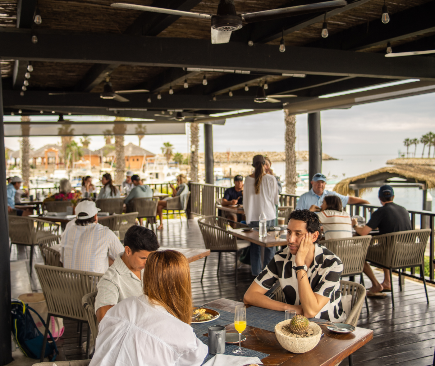 Outdoor restaurant patio overlooking marina with people dining and socializing under a wooden ceiling with ceiling fans.