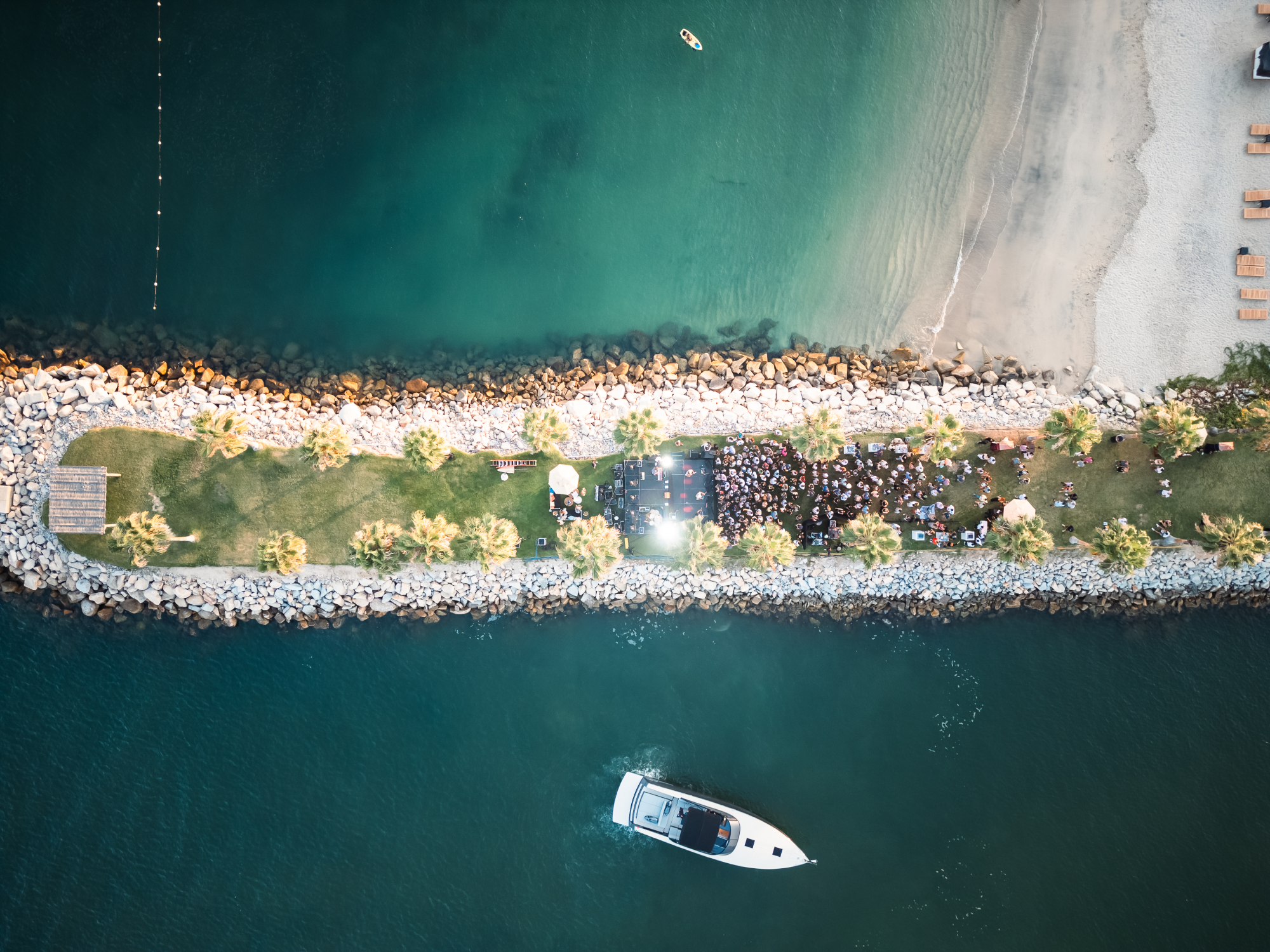 Aerial view of a narrow strip of land with palm trees and a large gathering of people, surrounded by water, with a boat nearby.