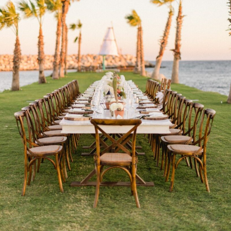 Long outdoor dining table with wooden chairs set on grass by the ocean, decorated with white flowers and glassware under palm trees at sunset.