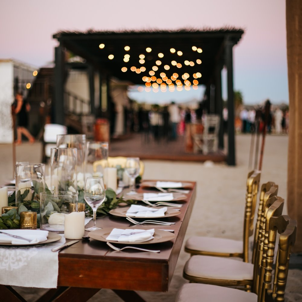 Elegant outdoor dining table set with plates, napkins, glassware, candles, and greenery, with a blurred pavilion and guests in the background.