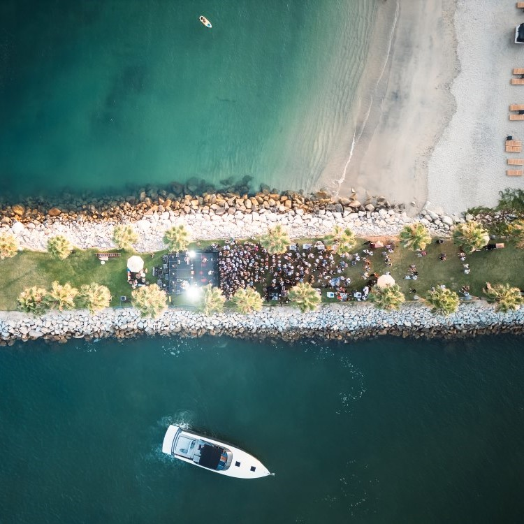 Aerial view of a narrow beach strip with green grass, palm trees, a crowd gathered in the center, a white boat in the water, and a small kayak near the shore.