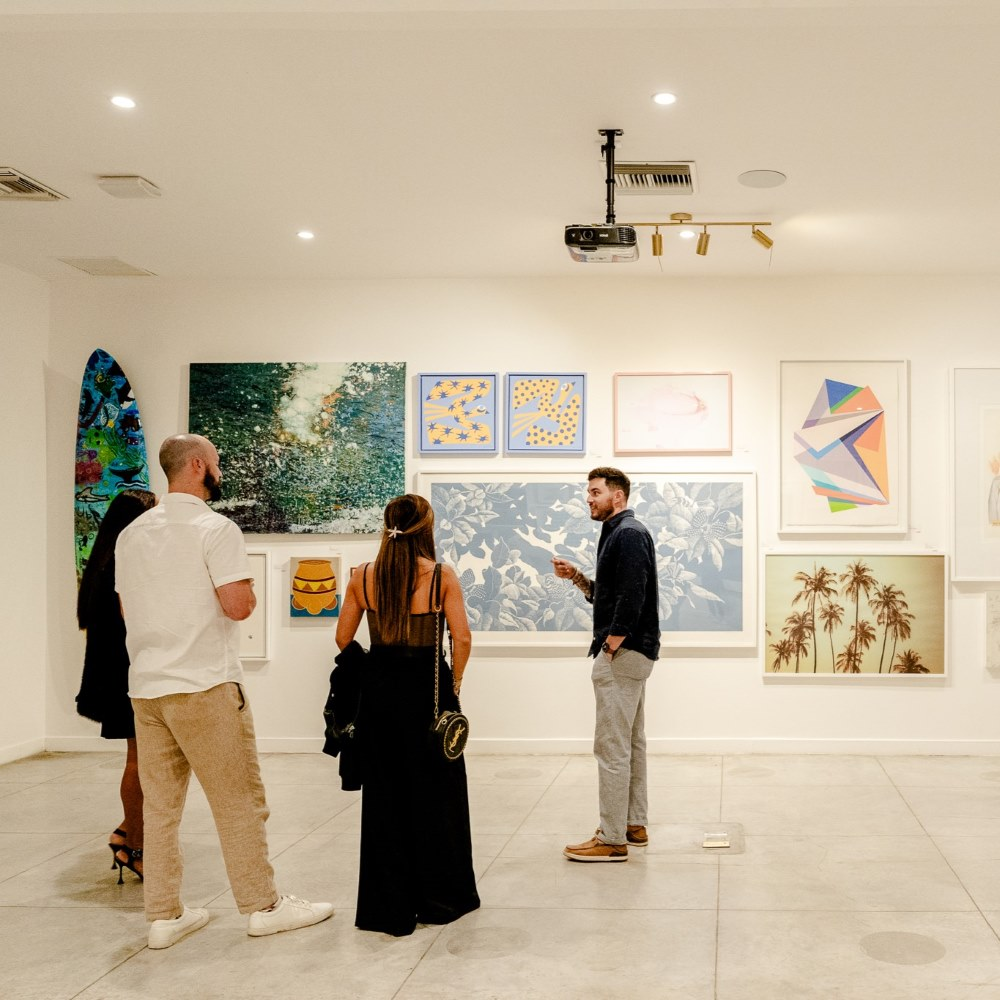 Four people in an art gallery observing and discussing various paintings and artworks displayed on a white wall.