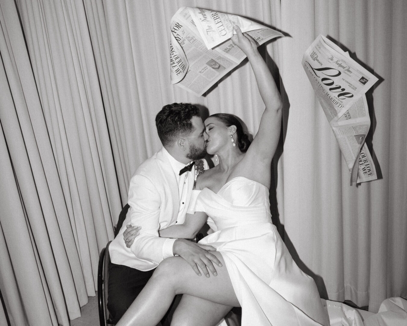 Bride and groom kissing, bride holding up newspapers with wedding announcements.