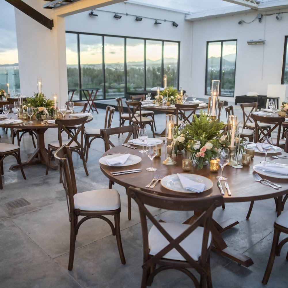 Indoor dining area with round wooden tables set with plates, napkins, glassware, and floral centerpieces with candles.