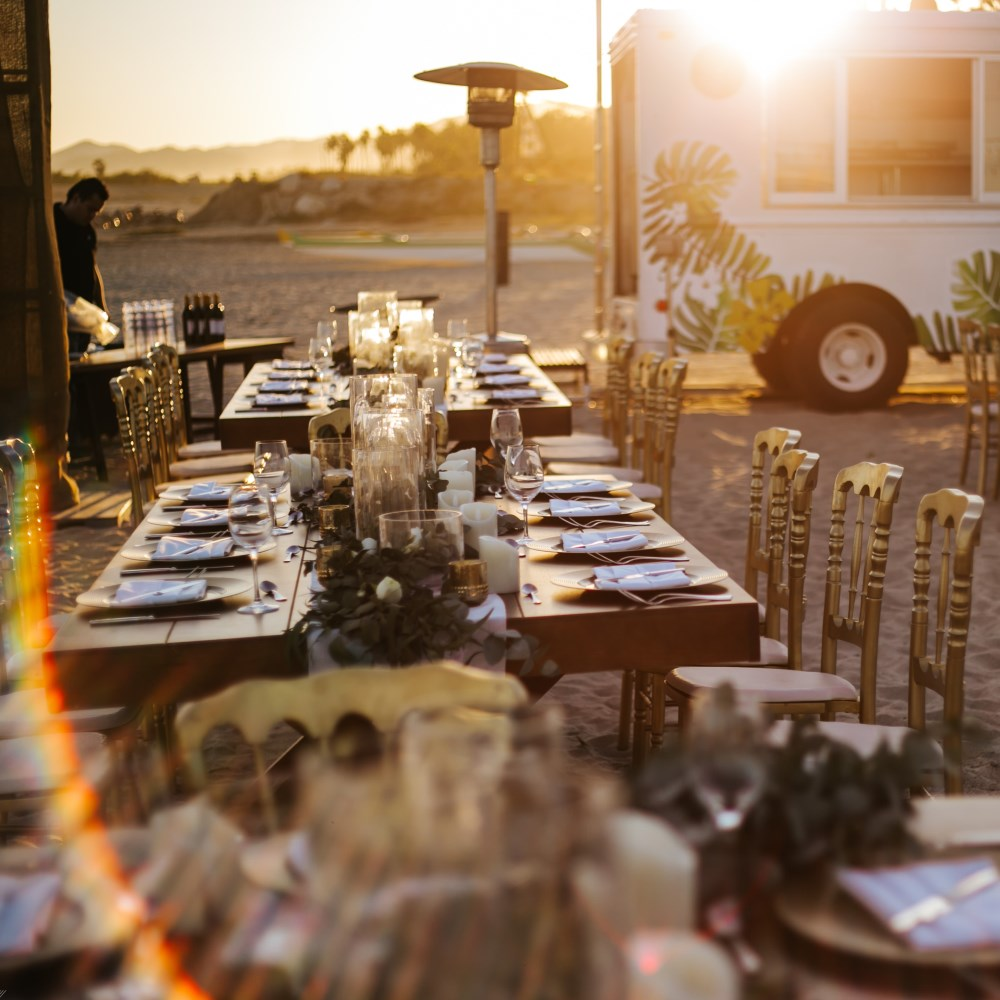 Elegant outdoor dining tables set with plates, napkins, glasses, and candles on a sandy beach at sunset.