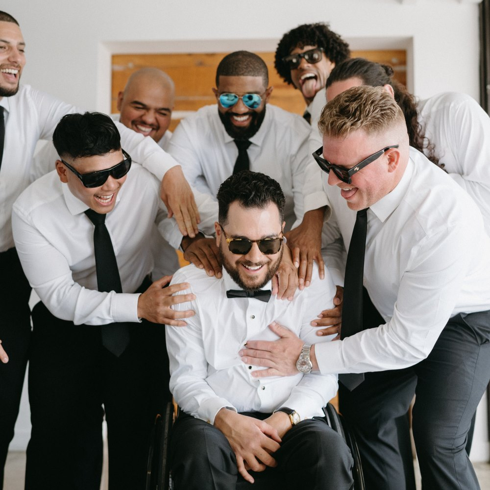 Group of men in white shirts and black ties, wearing sunglasses, smiling and surrounding a seated man in a wheelchair dressed in a white shirt and bow tie.