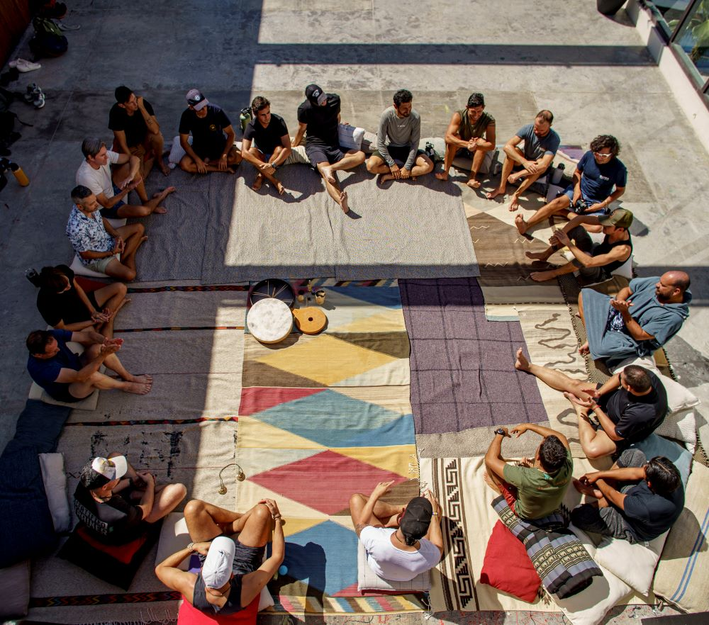 Group of people sitting in a large circle on colorful rugs and cushions outdoors, engaged in discussion or meditation.