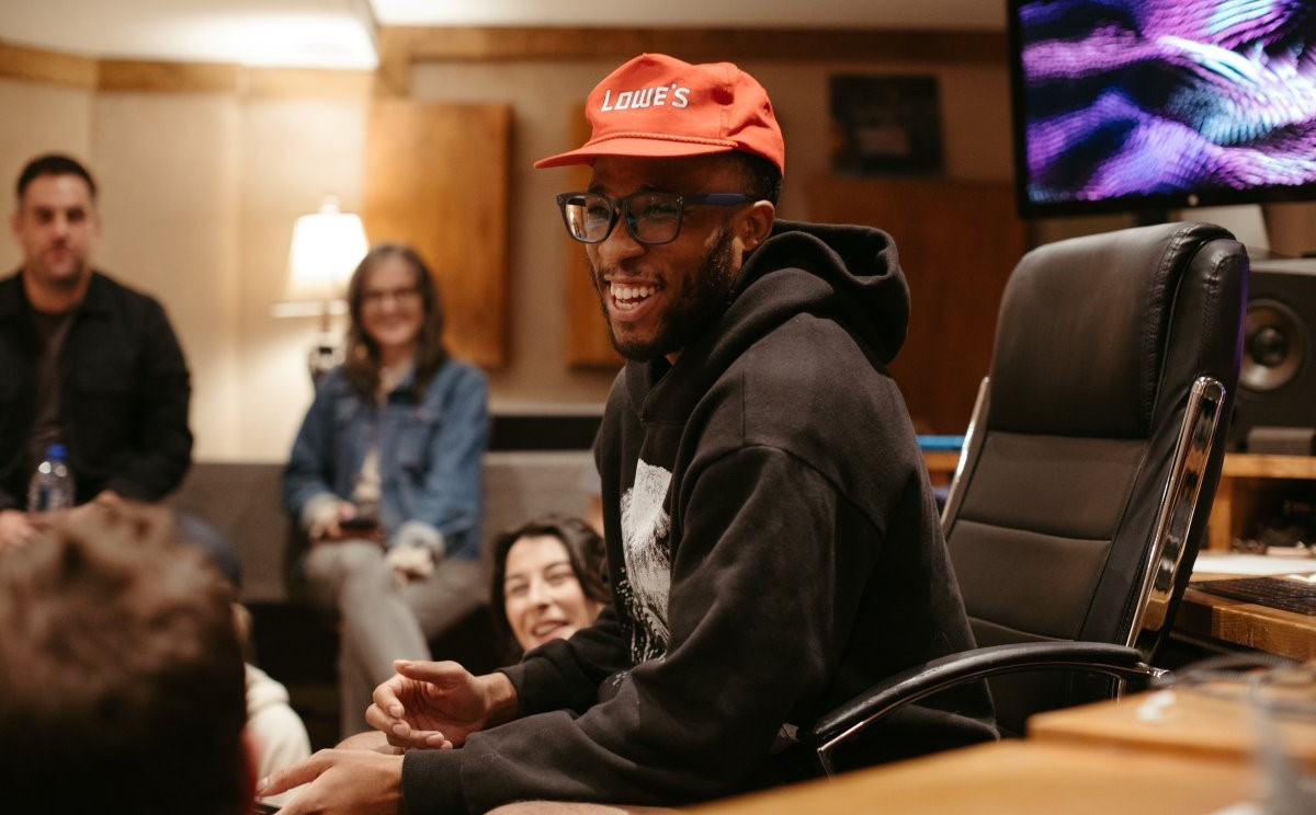 Man wearing glasses and a red Lowe's cap smiling and talking in a casual office setting with other people seated around him.