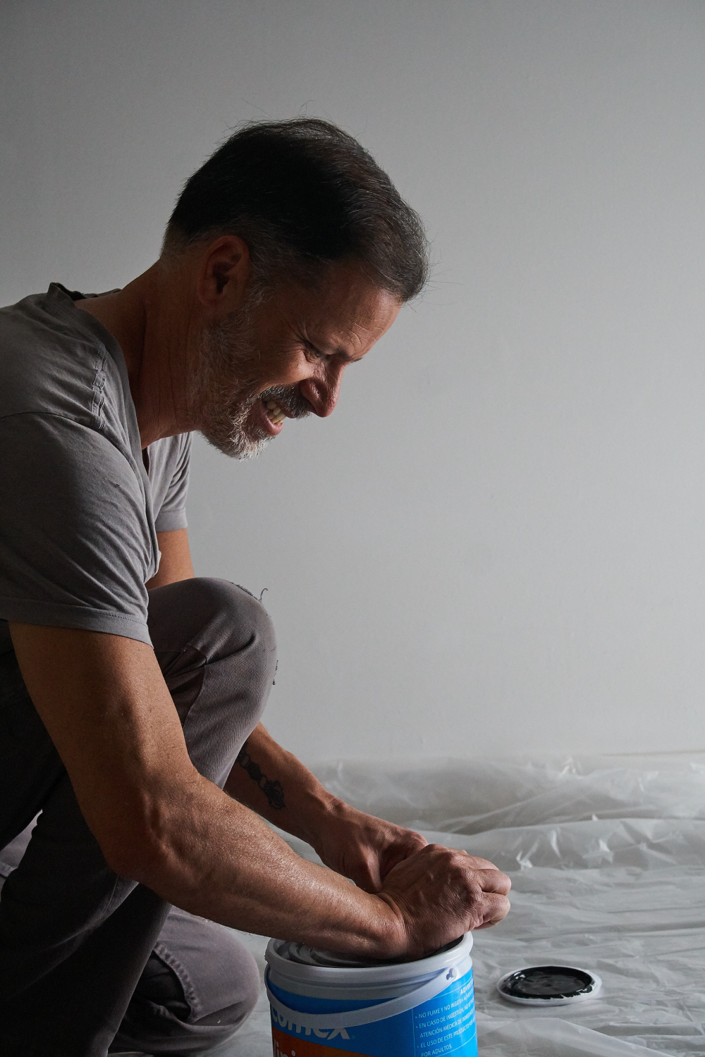 Smiling man kneeling and opening a white bucket with a blue label on a plastic-covered floor.